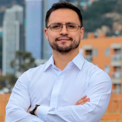 Photo of Julián Castro, Colombian man, wearing glasses, smiling, arms folded, city buildings and mountain in background